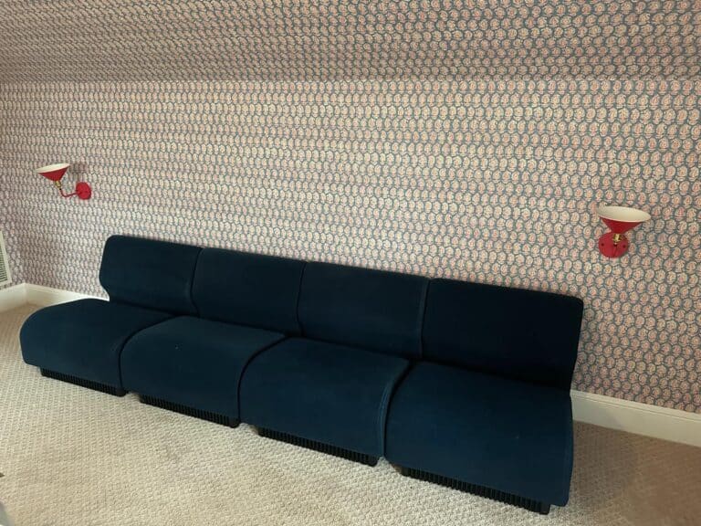 Room with a Herman Miller Blue Sectional, patterned wallpaper, and two red wall-mounted lamps.