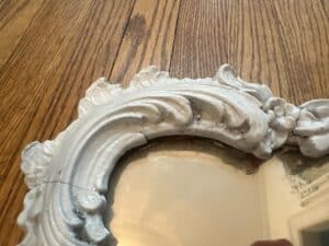 A close-up reveals the vintage white ornate frame of a Vintage Chinoiserie Mirror, showcasing visible cracks as it rests on a wooden surface.