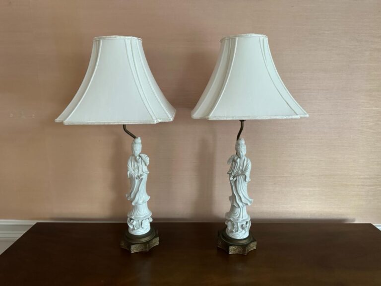 A pair of Blanc De Chine Figural Lamps with fabric shades sits on a wooden surface against a beige wall.