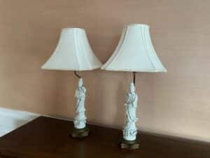 A pair of Blanc De Chine Figural Lamps with fabric lampshades sits on a wooden surface against a beige wall.
