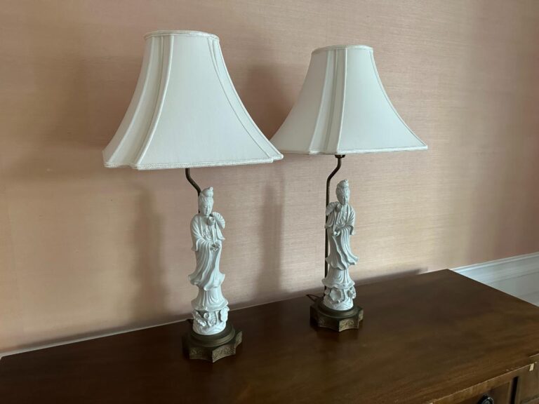 A pair of Blanc De Chine Figural Lamps with white shades and ornate bases placed on a wooden surface.
