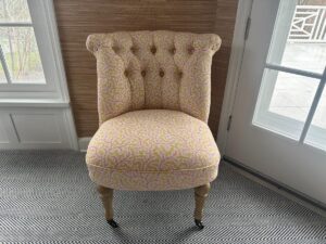 The Petite Tufted Back Side Chair on Casters, featuring a pink and yellow pattern, stands on a herringbone-patterned floor near a window.