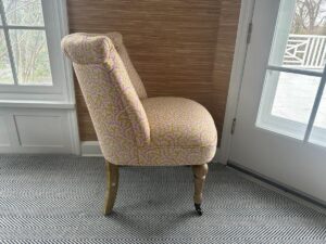 A side view of the Petite Tufted Back Side Chair on Casters with a patterned upholstery and wooden legs, placed on a herringbone carpet near a window and door.
