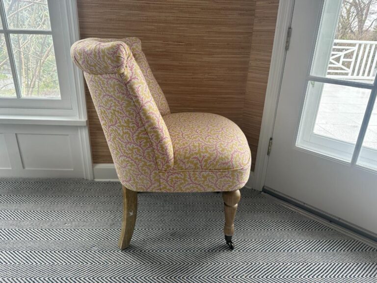 A side view of the Petite Tufted Back Side Chair on Casters with a patterned upholstery and wooden legs, placed on a herringbone carpet near a window and door.
