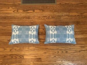 A pair of Sister Parish Mahalo Print Pillows featuring a blue pattern lies on a hardwood floor beside a floor vent.