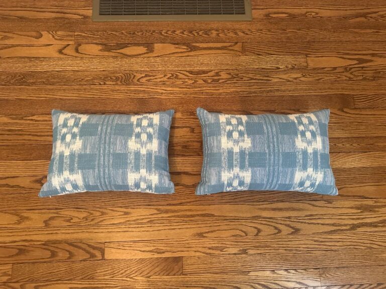 A pair of Sister Parish Mahalo Print Pillows featuring a blue pattern lies on a hardwood floor beside a floor vent.