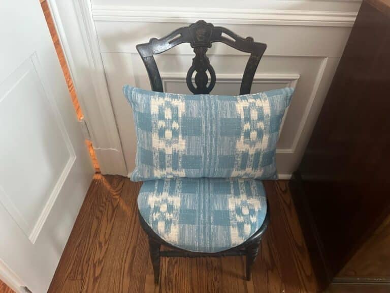 A pair of Sister Parish Mahalo Print Pillows with an ornate pattern rests on a wooden chair, which is set against a white-paneled wall on a wooden floor.