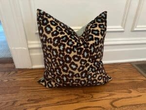 The Cut Velvet Leopard Pillow rests on a wooden floor, propped against a white-paneled wall.