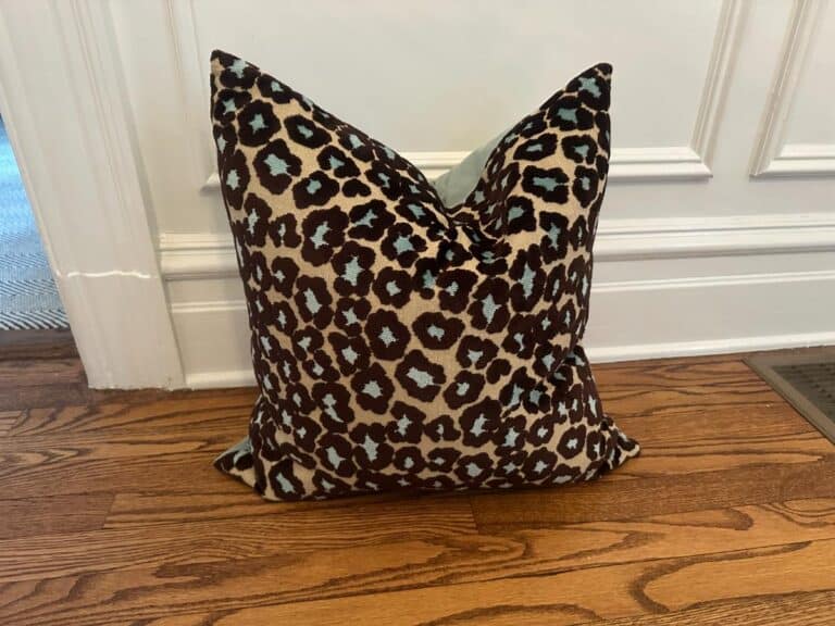 The Cut Velvet Leopard Pillow rests on a wooden floor, propped against a white-paneled wall.