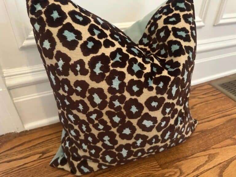 The Cut Velvet Leopard Pillow, featuring brown and turquoise spots, rests on a wooden floor near a vent.