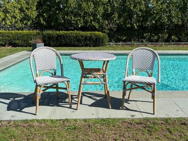 Tommy Hilfiger Blue & White Bistro Set on stone patio features two woven chairs and a round table, encircled by greenery.