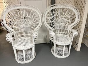 A pair of Vintage Hollywood Regency Style Peacock Chairs, showcasing intricate white wicker patterns, stand against a white wall.