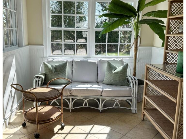 A sunlit room features a white wicker sofa with green cushions, a potted plant, large windows, and a Vintage Rattan Bar Cart.