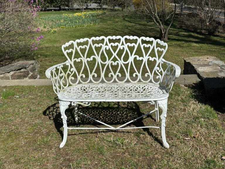 A White Cast Iron Garden Bench with heart motifs rests on a grassy lawn, surrounded by trees and stone features.