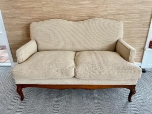 Chenille upholstered settee with wooden legs in front of a woven textured wall, on a zigzag patterned rug.