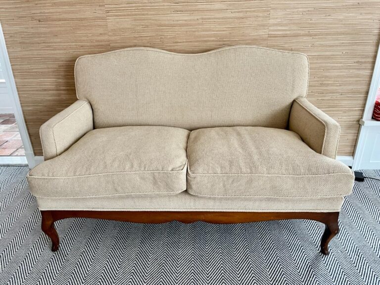 Chenille upholstered settee with wooden legs in front of a woven textured wall, on a zigzag patterned rug.