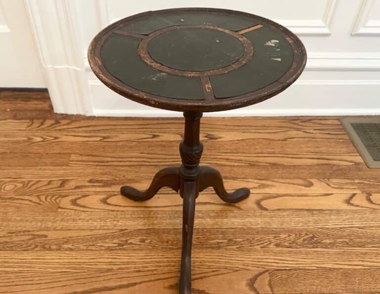 An Antique Side Table with a worn wooden round top, supported by a three-legged wooden base, sits on a wooden floor.