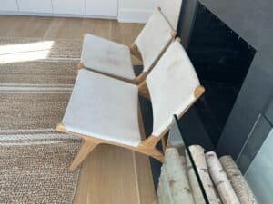 Two modern wooden chairs with white seats next to a fireplace, on a woven rug and wooden floor.