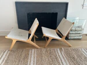 Two modern wooden lounge chairs with white cushions sit in front of a black fireplace and stacked firewood.