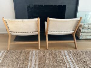 Two light wooden chairs with white cushions face a fireplace, next to stacked firewood on a glass holder.