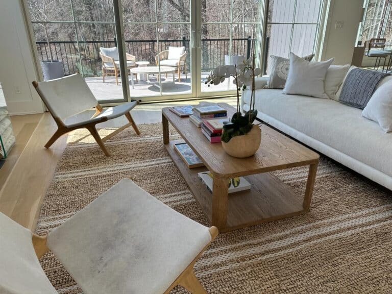 Modern living room with a woven rug, wooden coffee table, white chairs, and a sofa near large windows.