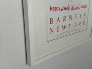 Framed poster on a white wall with red text reading "BARNEYS NEW YORK" and "Happy and Warm-lidays!.