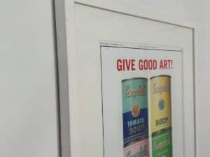 Framed poster with Campbell’s soup cans and the words “GIVE GOOD ART!” displayed on a white wall.