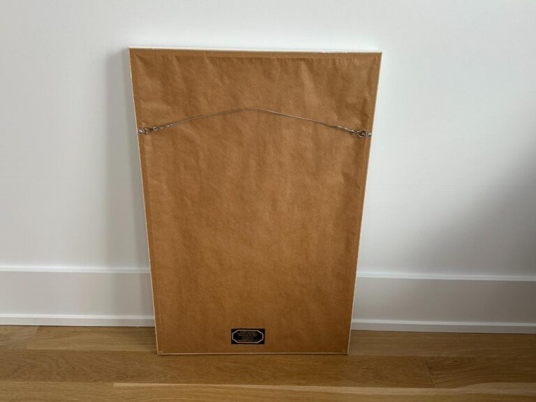 A framed picture turned backward, showing brown paper backing and a hanging wire, resting on a wooden floor.