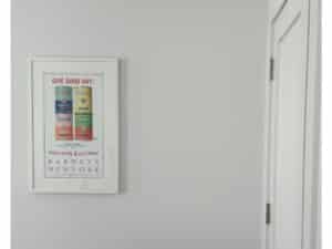 Framed poster of Campbell’s soup cans on a white wall next to a closed white door.
