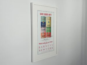 Framed poster of colorful soup cans with "GIVE GOOD ART!" and "BARNEYS NEW YORK" on a white wall.