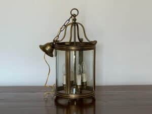 A brass and glass hanging lantern with three candle-shaped bulbs sits on a wooden table against a plain wall.
