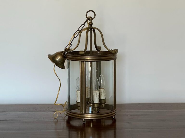 A brass and glass hanging lantern with three candle-shaped bulbs sits on a wooden table against a plain wall.