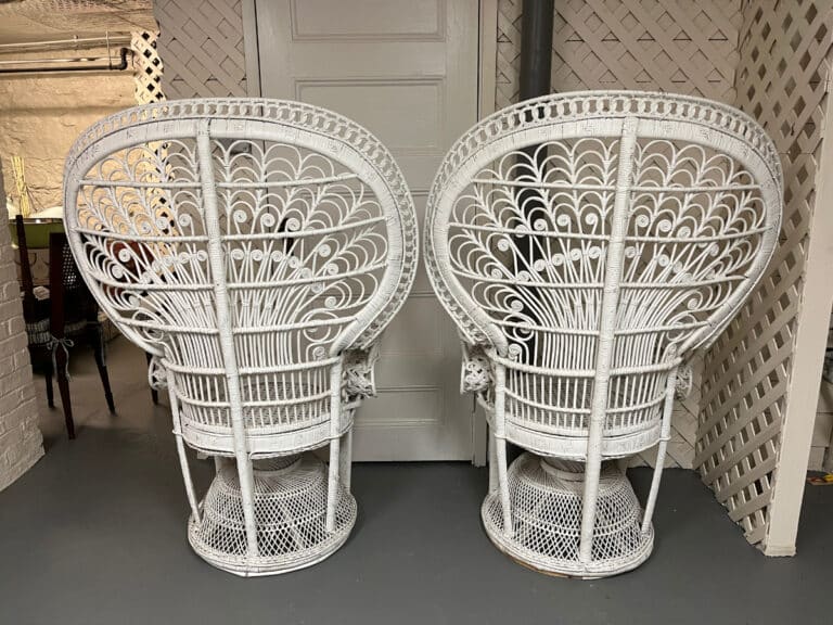 Two Vintage Hollywood Regency Style Peacock Chairs with ornate white wicker and tall, fan-shaped backs sit on a gray floor against a white lattice wall.