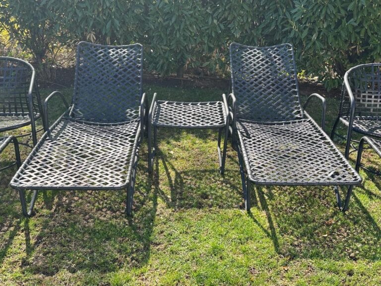 Two metal lounge chairs with a small table between them on grass, surrounded by a hedge.