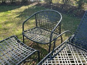 Metal patio furniture with a woven pattern on grass, surrounded by bushes under sunlight.