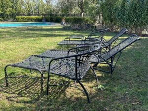 Four metal lounge chairs on grass, near a pool, surrounded by trees and a fence in the background.