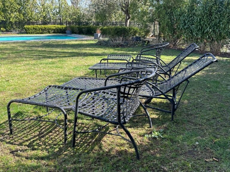 Four metal lounge chairs on grass, near a pool, surrounded by trees and a fence in the background.