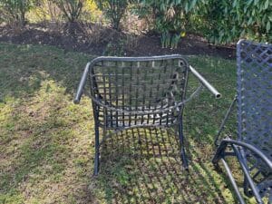Outdoor metal chair with armrests missing, on grass with bushes in the background.