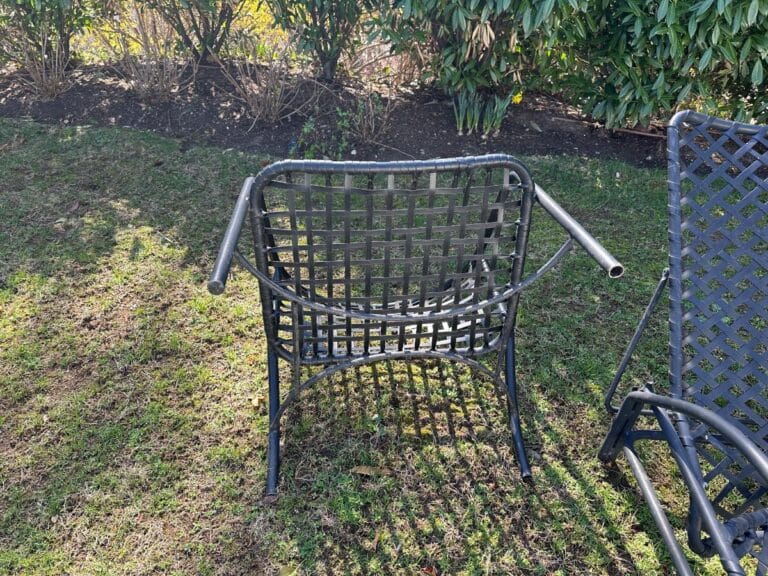 Outdoor metal chair with armrests missing, on grass with bushes in the background.