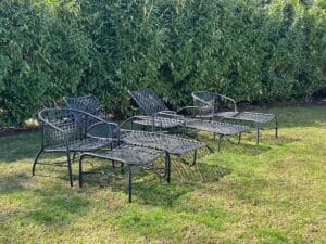 Four metal lounge chairs with footrests on green grass, set against a backdrop of tall, dense shrubbery.