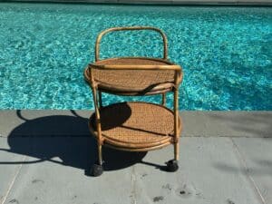 Rattan bar cart with two tiers on wheels, placed by a shimmering blue swimming pool.