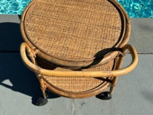 Wicker rolling cart with two shelves, standing on stone tiles near a swimming pool.