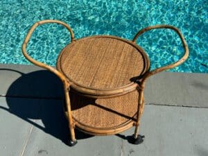 A two-tiered wicker rolling cart is positioned beside a pool on a sunny day.