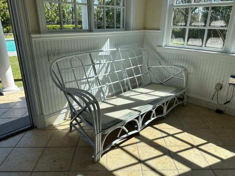 White wicker sofa with light cushions in a sunlit room, near windows overlooking a yard with a pool.