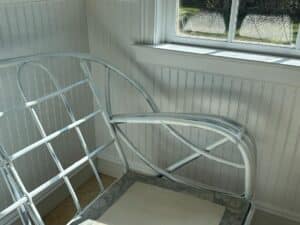 White metal chair frame in a sunlit room with a window, next to a white wooden paneled wall.