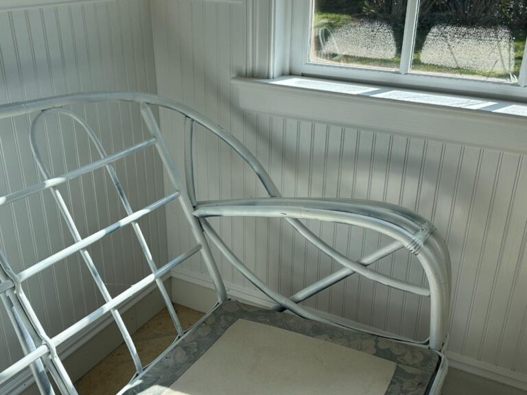 White metal chair frame in a sunlit room with a window, next to a white wooden paneled wall.