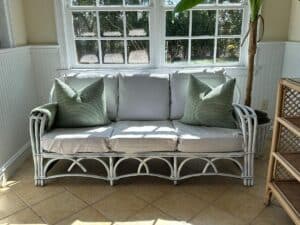 Wicker sofa with white cushions and green pillows in a sunlit room by a window and a potted plant.