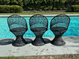 Three black wicker chairs on a patio by a pool, with greenery in the background.