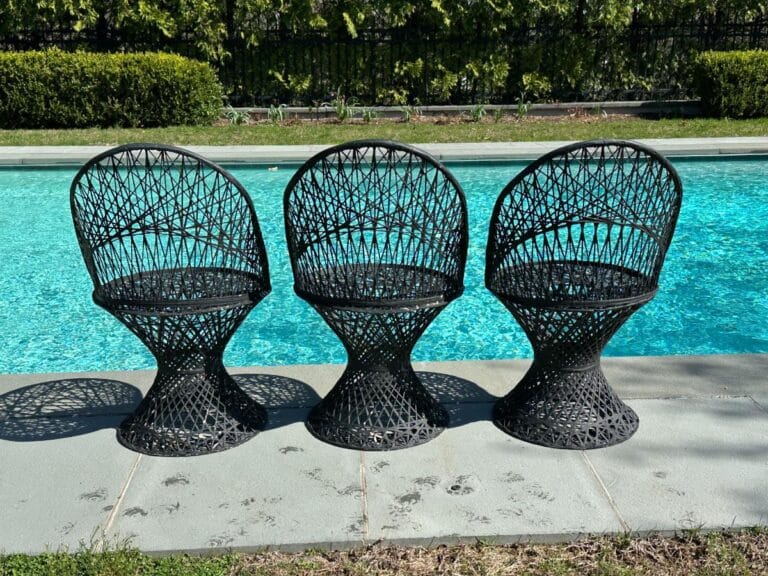 Three black wicker chairs on a patio by a pool, with greenery in the background.