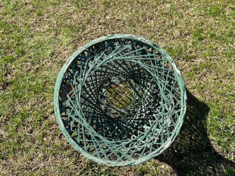 A round, green metal basket on grass, showing a complex crisscross pattern from above.